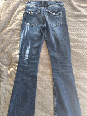 Mörkblå bootcut jeans - Säljer ett par mörkblå bootcut jeans med slitna detaljer och rå kant nedtill, då dom är uppsprättade. Jeansen har klassiska fem fickor, kontrastsömmar och en lätt tvättad look. 