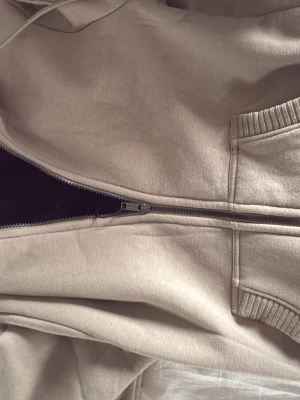 Beige zip-hoodie från DKNY - Snygg beige hoodie från DKNY med dragkedja framtill och två stora fickor. Hoodien har ribbade muddar vid ärmslut och nederkant samt en klassisk huva med dragsko. Tillverkad i mjukt sweatshirtmaterial som är skönt att bära.