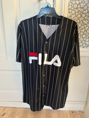 Svart randig skjorta/tshirt från FILA - Snygg svart skjorta/tshirt från FILA med vita vertikala ränder och stor logga framtill. Skjortan har knappar längs hela framsidan och korta ärmar. Perfekt för en sportig och avslappnad stil.