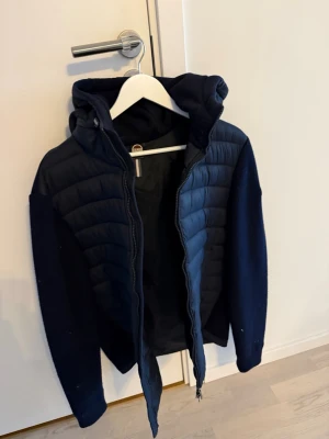 Mörkblå colmar jacka - Snygg mörkblå cardiganjacka med vadderad front och rygg samt stickade ärmar och sidor. Jackan har huva, dragkedja framtill och en sportig look. Herr jacka storlek 52/ L