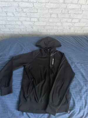 Svart Nike Running hoodie - Snygg svart hoodie från Nike Running med dragkedja framtill och huva. Jackan har en bröstficka med dragkedja och reflekterande Nike Running-tryck samt två sidofickor med dragkedja. Perfekt för löpning eller träning utomhus.
