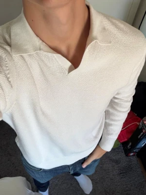 Vit stickad tröja med krage - Säljer en stilren vit stickad tröja med krage och v-ringad öppning. Tröjan har lång ärm och är tillverkad i ett mjukt, strukturerat material som ger en modern look. Perfekt att matcha med jeans för en clean outfit.