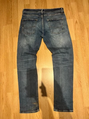 Blå jack & jones jeans - Säljer ett par klassiska blå jeans med raka ben och snygga slitningar. Jeansen har två bakfickor med dekorativa sömmar och normalhög midja. Materialet är denim i bomull och färgen är tvättad blå med ljusare partier.