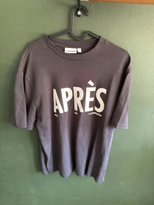 Grå t-shirt med tryck från J.Lindeberg - Säljer en grå t-shirt från J.Lindeberg med stort vitt tryck 'APRÈS' på bröstet. Klassisk rund hals och korta ärmar. Tillverkad i mjuk bomull för skön känsla och enkel stil.