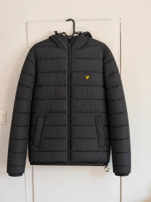Lyle & Scott Pufferjacka Svart – Strl M 🖤 - Snygg vadderad jacka från Lyle & Scott i svart/mörkgrå färg. Klassisk modell med huva, dragkedja framtill, två sidofickor och den gula Lyle & Scott-loggan på bröstet. Storlek: M Färg: Svart / mörkgrå Skick: Bra begagnat skick men jag har fixat en skada som man kan se på bild 5!😁 Detaljer: Vadderad modell, huva och sidofickor. En stilren och lättburen jacka som passar perfekt till vardags under höst, vinter och vår. Hör gärna av dig vid frågor eller om du vill ha mått! 📏