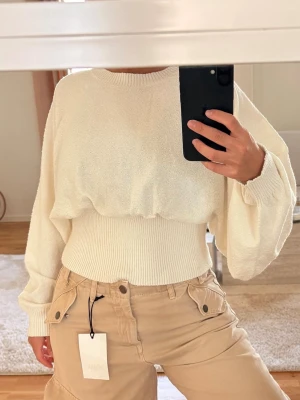 Vit croppad stickad tröja - Trendig vit stickad tröja med croppad passform och bred ribbad midja. Tröjan har ballongärmar och rund halsringning. Perfekt för dig som gillar en avslappnad men ändå stilren look. Materialet ser mjukt och bekvämt ut.