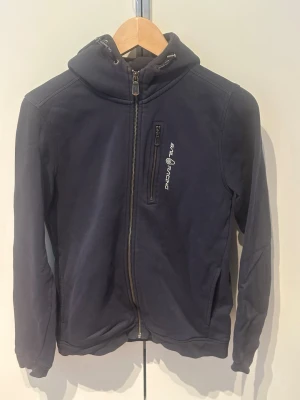 Marinblå hoodie från Sail Racing - Marinblå hoodie från Sail Racing med dragkedja framtill och bröstficka med logga. Huva med dragsko och metalländar. 