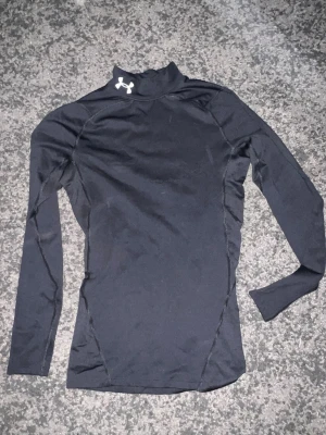 Svart långärmad träningströja UA - Säljer en svart långärmad träningströja från Under Armour med hög krage och tight passform. Tröjan har vit UA-logga vid halsen och är tillverkad i ett stretchigt funktionsmaterial som passar perfekt till träning eller löpning.
