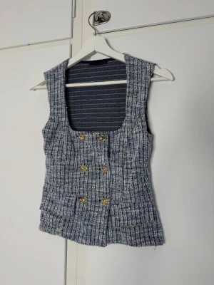 Blåvitt tweed set med guldknappar - Tvådelat set i blåvitt tweedmaterial med strukturerad känsla. Överdelen är ärmlös med breda axelband, fyrkantig urringning och dubbla rader guldknappar framtill. Kjolen är kort och matchar toppen perfekt. Perfekt för dig som gillar trendiga och stilrena plagg.