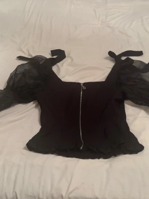 Helt ny Svart topp med puffärmar och dragkedja, boohoo - Snygg svart topp med transparenta puffärmar och knyt på axlarna. Toppen har en markerad midja och dragkedja framtill som detalj. Perfekt för dig som vill sticka ut lite extra.
