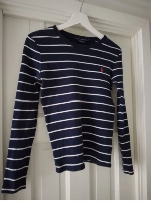 Ralph Lauren Sport mörkblå, vit långärmad tröja / topp, storlek S - En långärmad tröja från Ralph Lauren Sport. Tröjan har ett klassiskt randigt mönster i mörkblått och vitt, med den ikoniska polospelarlogotypen broderad i rött på bröstet. Den har en rund halsringning och en smal passform. Storlek: Small Skick: Bättre begagnad, inga defekter. Material: Bomull.