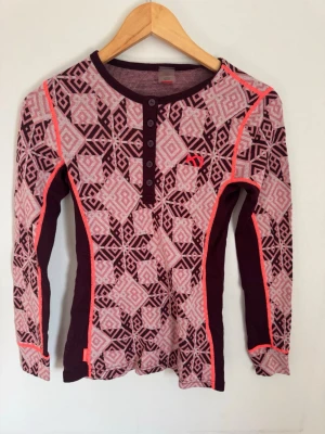 Mönstrad underställströja Kari Traa - Underställströja från Kari Traa i vinrött och rosa med geometriskt mönster. Långärmad modell med knappar vid halsen och kontrasterande sömmar i neonrosa. Perfekt för träning eller skidåkning. Materialet känns mjukt och stretchigt.