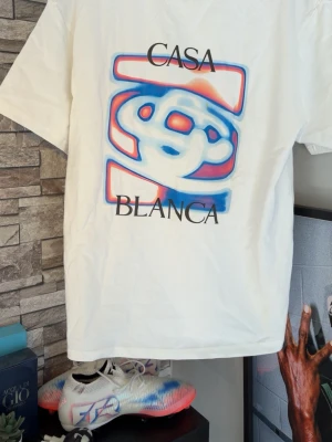 Vit Casa Blanca t-shirt med tryck - Vit t-shirt med färgstarkt grafiskt tryck i blått och rött samt texten 'Casa Blanca' på både bröstet och ryggen. Klassisk passform och rund halsringning. Perfekt för dig som gillar streetwear och unika prints.