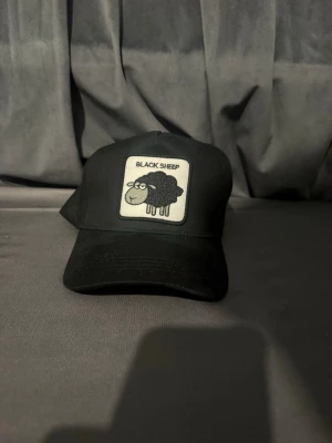 Svart keps med Black Sheep-tryck - Säljer en svart keps med böjd skärm och ett tygmärke framtill med texten 'BLACK SHEEP' och en tecknad svart får. Kepsen har justerbar snapback baktill och är tillverkad i ett klassiskt bomullsmaterial. Perfekt för dig som vill sticka ut med en rolig detalj.