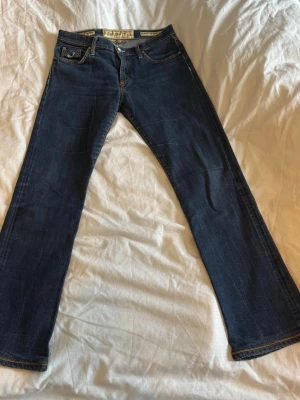 Mörkblå bootcut jeans - Snygga mörkblå bootcut jeans med klassiska fem fickor och kontrastsömmar. Jeansen har låg midja och detaljerade sömmar vid fickorna. Perfekta för en avslappnad och trendig look. Storlek w31 men sitter mer tajtare.