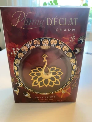 Plume D'Éclat Charm parfym - Plume D'Éclat Charm är en Eau de Toilette för kvinnor med en lyxig förpackning i vinrött och guld. Flaskan har en unik design med en vit och guldig påfågel i mitten, omgiven av ett dekorativt mönster. Parfymen rymmer 100 ml och har en elegant och feminin känsla.