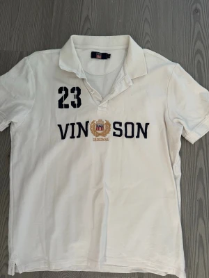 Vit pikétröja från Vinson - Vit pikétröja från Vinson med klassisk krage och knappar vid halsen. Tryck med siffran 23 och broderad logga samt texten 'VINSON ORIGINAL' på bröstet. Skön och avslappnad passform, perfekt till jeans eller shorts.