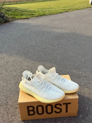 Yeezy Boost 350 V2 Cream White - Säljer ett par Yeezy Boost 350 V2 Cream White med stickad ovandel i vitt och en chunky, ribbad sula i ljusgul. Skorna har klassisk siluett, rund tå och vita skosnören. Perfekta för dig som gillar streetwear och vill ha något som sticker ut.