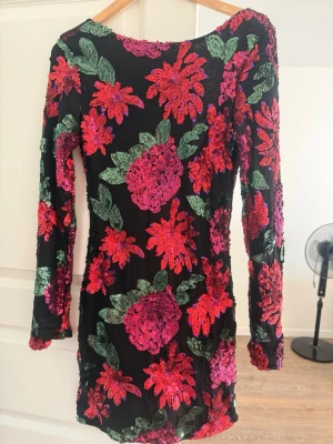 Svart paljettklänning med blommor - Kort svart klänning täckt av färgglada paljetter i rött, rosa och grönt som bildar ett blommigt mönster. Klänningen har lång ärm och en djup, öppen rygg. Perfekt för dig som vill sticka ut med en glittrig look.
