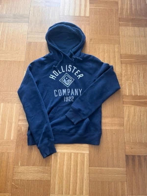 Mörkblå hoodie från Hollister - Mörkblå hoodie från Hollister med tryck 'Hollister Company 1922' i vitt på bröstet. Klassisk huva med dragsko och mjukt, fleecefodrat material. Ribbstickade muddar vid ärmslut och nederkant. Perfekt för en avslappnad och stilren look.