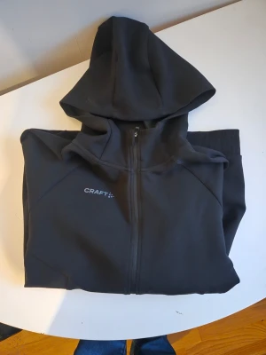 Svart hoodie med dragkedja från Craft - Snygg svart hoodie från Craft med hel dragkedja framtill och två fickor med dragkedja. Tröjan har huva och ribbade muddar vid ärmsluten. Perfekt för en sportig och avslappnad stil. Materialet känns mjukt och funktionellt.