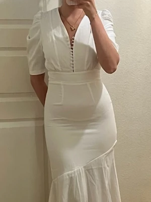 Vit långklänning med puffärm - Elegant vit långklänning med v-ringning och knappar framtill. Klänningen har puffärmar, markerad midja och en asymmetrisk volangkant nedtill. Materialet ser ut att vara mjukt och följsamt, perfekt för sommarens tillfällen.