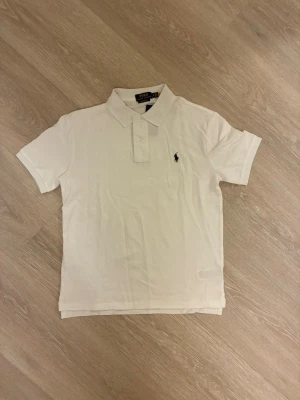 Vit pikétröja från Ralph Lauren - Klassisk vit pikétröja från Ralph Lauren med korta ärmar och krage. Tröjan har två knappar framtill och den ikoniska broderade loggan på bröstet. Tillverkad i bomull för en bekväm känsla och stilren look.