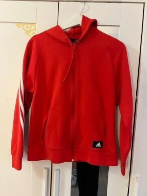 Röd Adidas hoodie med dragkedja - Röd hoodie från Adidas med dragkedja framtill och klassiska vita ränder längs ärmarna. Utrustad med huva och två fickor på sidorna. Adidas-logga på vänster sida nedtill. Perfekt för en sportig och avslappnad stil.