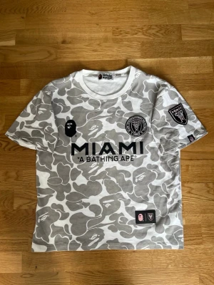 Grå camo BAPE x Miami T-shirt - Säljer en grå och vit camo t-shirt från BAPE i samarbete med Inter Miami. T-shirten har tryck fram och bak, med MIAMI-logga och BAPE 93 på ryggen. Klassisk rund hals och korta ärmar. Perfekt för dig som gillar streetwear och unika samarbeten.