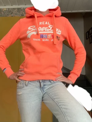 Vintage Superdry korall hoodie med tryck - Vintage hoodie i korall.Snygg orange hoodie från Superdry med vit och blå logga och japanska tecken på bröstet. Klassisk känguruficka framtill, ribbade muddar och dragsko i huvan. Perfekt för en avslappnad och trendig stil.