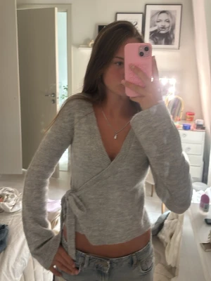 Grå omlottkofta från Gina Tricot - Mysig grå stickad omlottkofta från Gina Tricot med knytband i midjan. Koftan har långa ärmar och ribbade muddar vid ärmslut och nederkant. Perfekt för lager på lager och enkel att styla till olika outfits.
