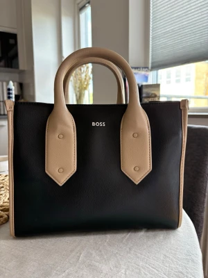 Svart och beige handväska från BOSS - Använd endast fåtal gånger. Jättebra skick men en liten skada på ena hörn, kolla bilder Nypris: 2385kr Mått: 21,0 x 26,5 x 13,0 centimeter. Priset kan diskuteras