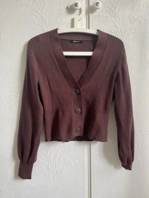 Brun ribbad kofta från Gina Tricot - Snygg brun ribbad kofta från Gina Tricot med djup v-ringning och stora knappar framtill. Modellen är croppad med långa ärmar och breda muddar. Perfekt att slänga över en topp för en chill look.