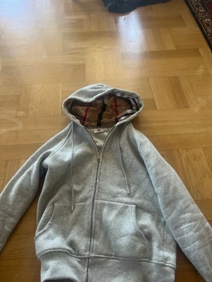Grå hoodie med mönstrad huva - Snygg grå zip-hoodie med dragsko och känguruficka. Insidan av huvan har ett klassiskt rutigt mönster i beige, rött och svart som ger en extra touch. Perfekt för en chill och avslappnad stil.