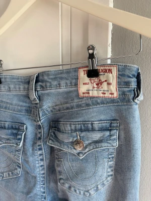 Ljusblå bootcut jeans True Religion - Säljer ett par ljusblå bootcut jeans från True Religion med ikoniska fickdetaljer och snygga kontrastsömmar. Jeansen har klassisk femficksmodell och silverfärgade knappar med logga. Perfekta för en avslappnad och trendig look. Storlek 28