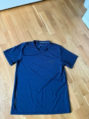 Under Armour t-shirt  - Tröjan har en liten fläck jämte loggan man kan se fläcken i bild 3.                                       Snygg blå tränings-tshirt från Under Armour med diskret logga på bröstet och tryck längs nederkanten. Tillverkad i ett lätt och snabbtorkande funktionsmaterial, perfekt för gymmet eller löprundan. Klassisk passform och rund hals.