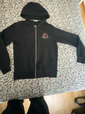 Svart Moncler hoodie med dragkedja - Säljer en svart hoodie från Moncler med dragkedja framtill och huva. På bröstet sitter Monclers ikoniska logga i rött och vitt. Hoodien har fickor framtill och ribbade muddar vid ärmar och nederkant. Perfekt för en chill och stilren look.