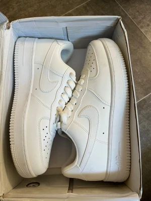 Nike Air Force 1 vita sneakers - Klassiska Nike Air Force 1 sneakers i helvitt läder med perforerade detaljer på tån och robust sula. Swoosh-logga på sidorna och Nike Air-brodyr på hälen. Perfekta för en clean och stilren look.