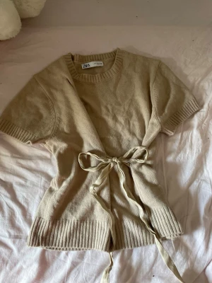 Beige stickad topp med knyt från Zara - Beige stickad topp från Zara med korta ärmar och snygg knytning i midjan. Toppen har ribbade detaljer vid ärmslut och nederkant, vilket ger en stilren och trendig look. Perfekt att matcha med jeans eller kjol.