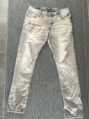Grå slim fit jeans med fem fickor från Jack and Jones - Snygga grå slim fit jeans med klassisk femficksdesign och dragkedjegylf. Jeansen har en tvättad look och kontrastsömmar, perfekta för en avslappnad stil. Materialet känns mjukt och stretchigt för extra komfort.