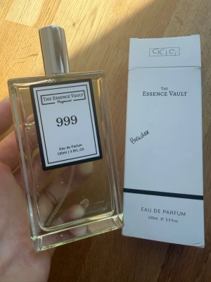 The Essence Vault 999 Eau de Parfum - Parfymen The Essence Vault 999 Eau de Parfum kommer i en stilren, rektangulär glasflaska med silverfärgad spraykork. Flaskan rymmer 100 ml och har en enkel, vit etikett med svart text. Förpackningen är vit med minimalistisk design. Doftar som Prada paradox