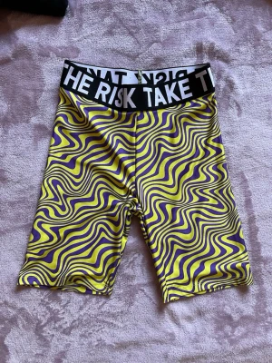 Gula och lila mönstrade cykelshorts, stl XS oanvända  - Färgstarka cykelshorts med psykedeliskt mönster i gult och lila. Bred svart resår i midjan med vit text 'TAKE THE RISK'. Tight passform och stretchigt material som passar perfekt till träning eller sport. Stl XS oanvända 