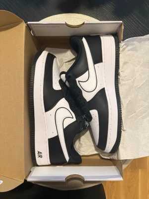 Nike Air Force 1 svart/vit sneakers - Säljer ett par Nike Air Force 1 sneakers i svart och vitt läder. Klassisk låg modell med svart yttersula, vita detaljer och svart swoosh-logga. Skorna har perforerad tåbox och svart snörning. Perfekta för dig som gillar stilrena och tidlösa sneakers. Bara testade hemma.