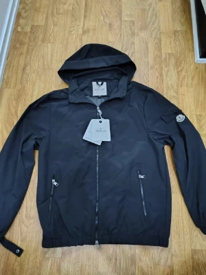 Svart vindjacka från Moncler - Snygg svart vindjacka från Moncler med huva och dragkedja framtill. Två fickor med dragkedja och Moncler-logga på ärmen. Jackan har elastisk nederkant och är tillverkad i ett lätt, vattenavvisande material. Perfekt för blåsiga dagar. Priset kan diskuteras!!!