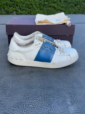 Valentino open blå - Snygga vita sneakers från Valentino med bred blå läderdetalj över snörningen. Klassisk låg modell med vita skosnören och diskret logga på plösen. Yttermaterial i läder och gummisula. Perfekta för dig som vill ha en stilren men ändå unik look. Storlek 42,5. Väldigt bra skick. Dustbag och box ingår. Enda dåliga är det ni kan se på bild 7. Men det är inget jag märker av och syns knappt. (Mina vänner har inte ens tänkt på det)  Pris kan diskuteras vid snabb affär! 🤝Självklart äkta. 