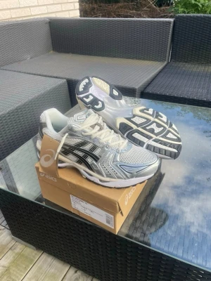 ASICS GEL-Kayano 14 silver sneakers - Snygga ASICS GEL-Kayano 14 sneakers i silver och vitt med svarta detaljer. Skorna har en sportig mesh- och syntetöverdel, klassisk snörning och en chunky sula med tydlig ASICS-logga på sidan. Perfekta för dig som gillar retrovibbar och streetstyle.