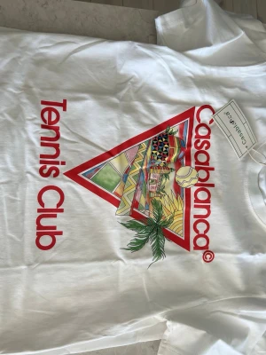 Vit Casablanca Tennis Club t-shirt - Säljer en vit t-shirt från Casablanca med färgglatt tryck och texten 'Tennis Club' i rött. T-shirten har klassisk passform och korta ärmar. Perfekt för dig som gillar sportig och trendig stil.