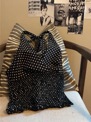 Svart prickig topp - Snygg svart omlottopp med vita prickar och knyt i nacken. Toppen har smockad nederdel och volangkant, vilket ger en feminin och trendig look. Perfekt för dig som gillar att sticka ut med detaljer och mönster.
