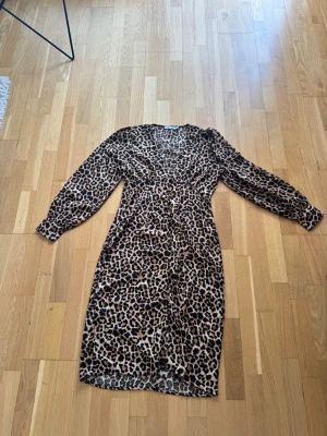 Leopardmönstrad klänning från NA-KD - Snygg leopardmönstrad klänning från NA-KD med v-ringning och knappar framtill. Klänningen har lång ärm och markerad midja som ger en smickrande siluett. Perfekt för dig som vill sticka ut med ett djurmönster.