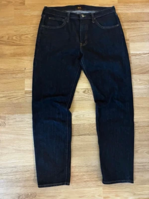 Mörkblå raka jeans från Lee - Säljer ett par mörkblå jeans från Lee med klassisk femficksmodell och raka ben. Jeansen har guldfärgade sömmar och knappgylf. Tillverkade i slitstark denim som ger en tidlös look. W33/L32